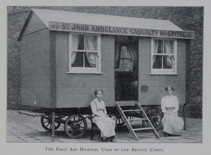 This is a black and white image of a mobile hospital. It is a small wooden room with four walls and a roof, and it is set on four wheels so that it can be transported. A four-step wooden ladder leads up to the entrance, which is flanked by two curtained windows on either side. Two women in light dresses are sitting either side of the ladder, in front of the hospital.