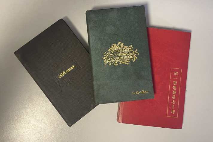 A photograph of three small hardback books lying on a table. The book on the left has a black cover, the book in the middle has a blue cover, and the book on the right has a red cover. Each book has a title embossed into the cover in gold.