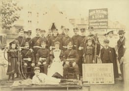 A black and white photograph of a group of men, women, and children standing and seated for a posed photograph. Sixteen of the men and one woman are in St John Ambulance Brigade uniform, and the remainder are in civilian dress. The group are outside in front of pointed tent, with a sign identifying it as a first aid tent. A boy is reclining in front of the group on a stretcher.
