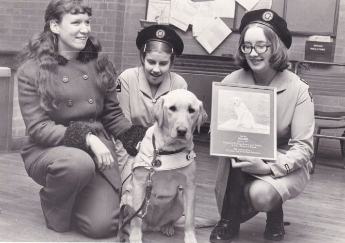 A black and white photograph showing three female Cadets kneeling in a row. Two of the Cadets are wearing St John Ambulance Cadet uniform, and the other is in civilian dress. The Cadet in civilian dress is blind, and in front of the three Cadets is her golden retriever guide dog in a harness. The Cadet on the right is holding a framed letter in thanks for raising funds for the Guide Dogs for the Blind Association.