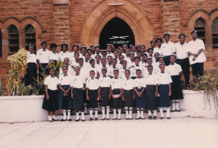 A colour photograph showing a group of 54 men, women, and children, wearing black and white Ghanaian St John Ambulance uniform and standing in rows on steps in front of a church. 