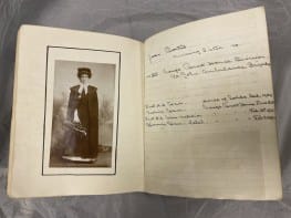 A photograph of a small lined notebook lying open on a grey cushion. On the left page is a black and white photograph of a female nurse wearing the uniform of the St John Ambulance Brigade. On the right page are handwritten notes.