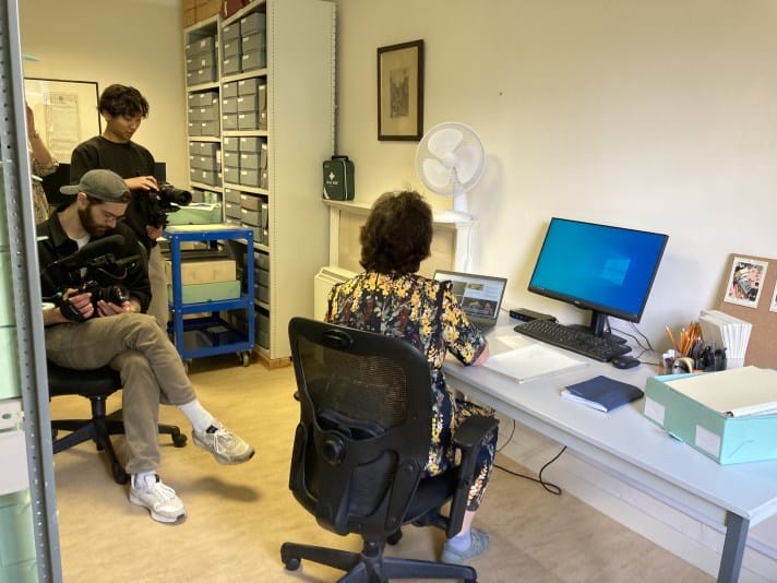 A view of a woman being filmed in her workplace. The woman has her back to the image viewer; she is sitting down at a desk working on a laptop. She has short dark hair and she is wearing a floral dress. To her left are two men; one standing, one seated. The man who is standing has short dark hair and is holding a videocamera which is pointed at the woman. The man who is seated is wearing light trousers, a dark jumper, and wearing a cap on his head. He is making notes.