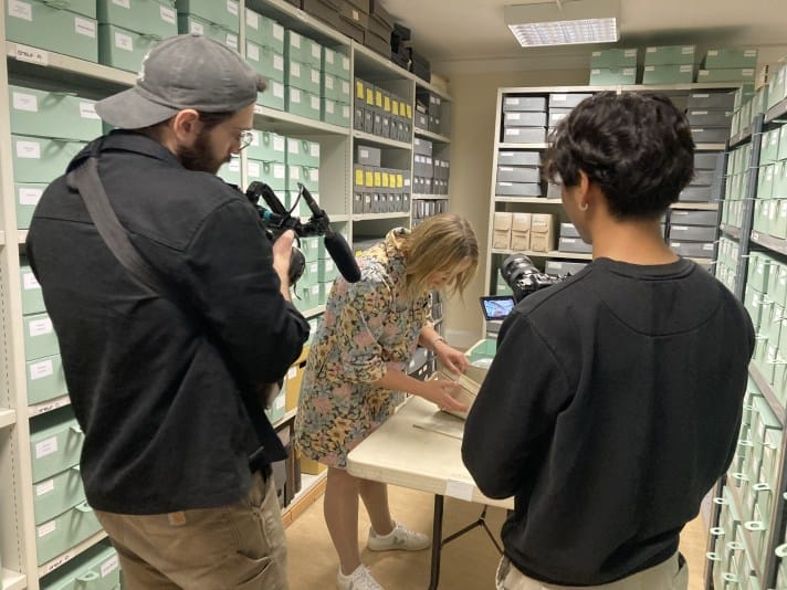 A view of a woman being filmed doing her work. In the centre is a woman with blonde hair in a knee-length light floral dress. She is rummaging through a green box which is sitting on top of a grey table. Behind here can be seen shelves filled with boxes. In the foreground are two men with their backs to the image viewer. They are both wearing dark jumpers and are holding cameras and microphones which they are pointing at the woman.