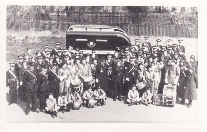 A black and white photograph from the 1930s of a large group of people, men, women, and boys, in St John Ambulance Brigade uniform, standing and sitting in front of a dark coloured ambulance. Some of the boys are holding drums, as they are part of a St John band.