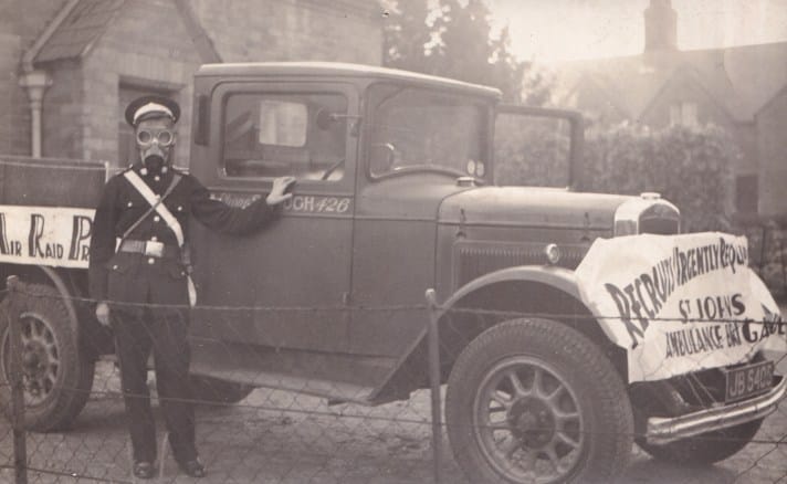 A black and white photograph from 1943 of a man wearing his St John Ambulance Brigade uniform and a gas mask, standing in front of a flatbed truck which has signs hanging from it to promote the St John Ambulance Brigade’s recruitment of Air Raid Precaution volunteers.