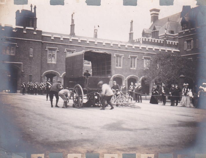 A black and white photograph from 1894 of three St John Ambulance Brigade volunteers demonstrating their first aid response to a staged incident of a person having been run over by a horse cart. The demonstration is watched by civilians and uniformed Brigade members, and is taking place at St. James’s Palace, which is visible in the background.