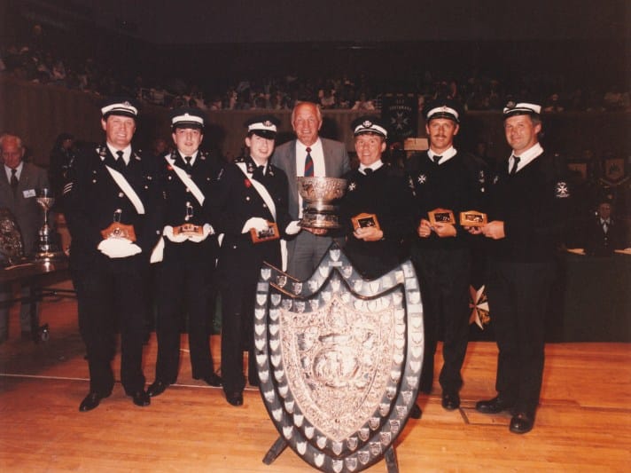 A colour photograph of seven men standing in a line. The person in the middle is Henry Cooper, a professional boxer, and the three men on either side of him are St John Ambulance volunteers wearing their uniforms. The volunteers are holding trophies and are also pictured with a large shield, all are prizes won in a first aid competition.