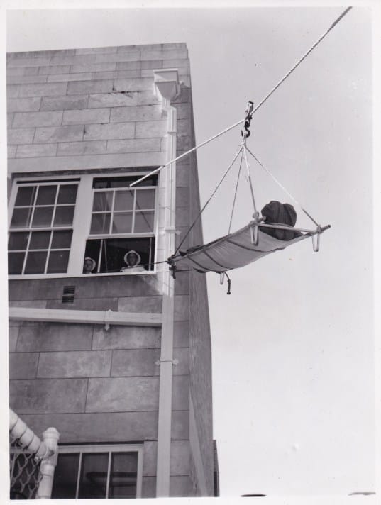 A black and white photograph of a simulated patient being stretchered out of a second-floor window using a winching system, while people look on.