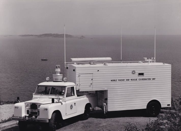 A black and white photograph of a Landrover which is towing a small portable office-style unit. It is parked at the top of a cliff, with the sea and an island visible in the background. On the unit are the words ‘Mobile Radar and Rescue Operation Unit’.