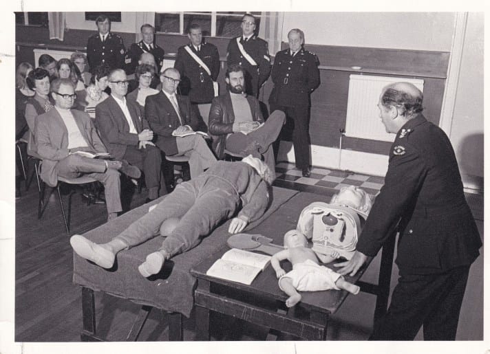 A black and white photograph of a first aid course taking place in a classroom. At the front of the room is a uniformed male St John Ambulance volunteer who is teaching the class, and in front of him are resuscitation aids. The are sitting down, facing him and listening attentively.