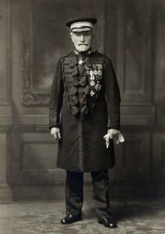 A black and white full-body portrait photograph of Sir James Cantlie. In the photograph, he is facine the camera and wearing his St John Ambulance Brigade ceremonial uniform and cap. On his left breast are his medals, and he is wearing the insignia of the Order of St John at his neck.