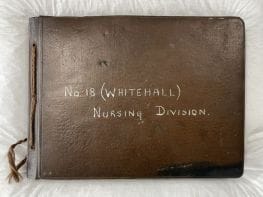 A colour photograph of a brown soft leather-backed photograph album lying on tissue paper. On the front and centre of the album are the words 'No. 18 (Whitehall) Nursing Division' handwritten in white pen.